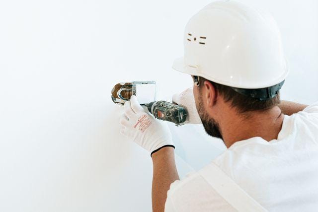 Contractor in a hard hat and gloves drilling into a white wall